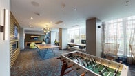 This dynamic amenity space is thoughtfully designed to offer a variety of recreational and functional zones within a single, open floor plan. The game area serves as a central hub, featuring a foosball table, a full-length shuffleboard table, and a ping-pong table set atop a boldly patterned carpet with teal and rust tones that lends energy to the space throughout.

A striking wood-slatted accent wall frames a built-in fireplace and wall-mounted flat-screen television, creating a warm, lounge-like focal point. Adjacent to this, a dedicated workstation zone lines the floor-to-ceiling windows, offering abundant natural light and urban views. Ergonomic seating, dual monitors, and a continuous desk surface make this area well-suited for focused work or casual browsing. A vibrant yellow pod chair adds a playful accent nearby.

A stylish chandelier with gold globe fixtures illuminates the central gathering area, complementing the contemporary interior palette of taupe, gold, and white. A glass-walled corridor leads to a private meeting room, visible through a frosted-glass partition, as well as restroom facilities. Framed artwork adorns the walls throughout, contributing to a polished yet approachable atmosphere. The overall design balances social engagement with functional workspaces in a cohesive, modern environment.