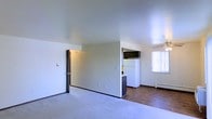 This single-floor unit features an open-concept layout that connects the main living area to the kitchen, creating a practical and cohesive floor plan. The living area is finished with wall-to-wall carpet and neutral white walls, lending the space a clean, understated aesthetic. A ceiling fan is visible toward the dining or transitional zone, providing both air circulation and a modest lighting fixture.

The kitchen is partially visible from the main living space and is separated by a partial wall, which adds subtle definition between areas without fully closing off the layout. The kitchen features a white refrigerator, dark upper cabinetry, and a raised countertop peninsula, transitioning onto hardwood-style flooring that contrasts with the carpeted living zone. A baseboard heater runs along the wall near the kitchen, indicating the unit's heating setup.

Natural light filters in through a window fitted with vertical blinds, brightening the right side of the space. A doorway on the left side of the main room leads to an additional area, likely a bedroom or hallway. Overall, the unit presents a functional, no-frills layout suited for everyday living.