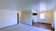 This single-floor unit features an open-concept layout that connects the main living area to the kitchen, creating a practical and cohesive floor plan. The living area is finished with wall-to-wall carpet and neutral white walls, lending the space a clean, understated aesthetic. A ceiling fan is visible toward the dining or transitional zone, providing both air circulation and a modest lighting fixture.

The kitchen is partially visible from the main living space and is separated by a partial wall, which adds subtle definition between areas without fully closing off the layout. The kitchen features a white refrigerator, dark upper cabinetry, and a raised countertop peninsula, transitioning onto hardwood-style flooring that contrasts with the carpeted living zone. A baseboard heater runs along the wall near the kitchen, indicating the unit's heating setup.

Natural light filters in through a window fitted with vertical blinds, brightening the right side of the space. A doorway on the left side of the main room leads to an additional area, likely a bedroom or hallway. Overall, the unit presents a functional, no-frills layout suited for everyday living.