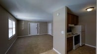 This single-floor space presents a practical and functional layout designed for comfortable everyday living. The open-concept design connects the living area and kitchen, creating a cohesive and accessible floor plan. The spacious living area is finished with wall-to-wall carpet in a neutral tone, complemented by warm beige walls and clean white baseboards that contribute to an understated, welcoming ambiance. A textured popcorn ceiling runs throughout, adding a classic residential character to the space. Natural light filters in through sidelights flanking the front entry door, brightening the living area. A secondary interior door provides access to additional areas of the unit. The kitchen is positioned adjacent to the living space, separated by a partial wall that defines each zone while maintaining an open feel. It features dark wood cabinetry offering ample storage, a white electric range with an overhead ventilation hood, and light-toned countertops. The kitchen floor transitions to tile, providing a durable and easy-to-clean surface in the cooking area. Overall, the space offers a straightforward and well-organized layout suited for practical, day-to-day living.