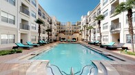 Located at 5555 E Mockingbird Ln in Dallas, Texas, this outdoor amenity space is designed to offer a resort-style experience within a multi-story residential community. At its heart is a generously sized swimming pool with crystal-clear blue water, framed by rows of turquoise lounge chairs and swaying palm trees that lend a tropical atmosphere under the open Dallas sky.

Adjacent to the pool, a beautifully appointed courtyard features classic brick paver flooring and multiple outdoor seating areas, each anchored by lush patches of turf. Rustic wood pergolas with string lighting serve as architectural focal points, providing shaded retreats ideal for relaxing or socializing. The pergola structures showcase exposed heavy-timber beams that add warmth and character to the otherwise modern, light-toned building facade surrounding the space.

The courtyard is enclosed by the multi-level building, whose balconies and metal railings overlook the amenity area, creating a sense of community and privacy simultaneously. Large glass-paned entry doors connect the outdoor space to interior common areas, ensuring seamless flow between inside and out. Decorative planters with lush greenery are thoughtfully placed throughout, adding natural vibrancy. Overall, the space strikes a balance between polished urban design and relaxed, outdoor comfort.