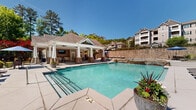 Located at 40 Chateau Dr SE in Rome, Georgia, this outdoor amenity space offers a resort-inspired pool complex designed for relaxation and recreation. The centerpiece is a generously sized swimming pool with clear, blue water, complemented by a separate hot tub with natural stone accents and a cascading water feature that adds a tranquil ambiance to the environment.

The expansive pool deck is finished in neutral concrete and features multiple lounge chairs and umbrella-shaded seating areas, providing ample space for sunbathing and socializing. A raised sun terrace, bordered by a striking stacked-stone retaining wall, offers additional seating with views overlooking the pool area.

A covered outdoor pavilion serves as the social hub of the space, anchored by stone-base columns supporting a white-painted roof structure. Ceiling fans ensure comfort during warmer months. The pavilion is equipped with a wall-mounted flat-screen television, a built-in serving counter with cabinetry, and multiple dining tables and chairs, making it ideal for gatherings. The surrounding landscape features mature trees and lush greenery, creating a naturally secluded setting. Iron fencing encloses the perimeter, adding both security and an elegant finish to this well-appointed outdoor retreat.