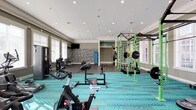 This fitness center offers a comprehensive and thoughtfully designed workout environment spread across a single floor. The space is immediately eye-catching, featuring a bold, teal-patterned carpet that energizes the room and defines the workout zones. Recessed lighting throughout the white ceiling ensures the entire area is well-lit, complementing the natural light that flows in through multiple windows fitted with blinds along the perimeter walls.

The layout accommodates a diverse range of exercise equipment, including Matrix spin bikes and cardio machines positioned toward the front of the space, along with a dedicated free weights area featuring a full dumbbell rack set against a decorative stone-textured accent wall. A striking green functional training rig with rope netting and barbell storage anchors one side of the room, catering to strength and functional fitness training. Additional resistance machines are positioned throughout, offering a variety of workout options.

The design elements add character to the space, with a teal accent wall, vertical wood-panel detailing, and an artistic stone wall mural creating visual interest. A wall-mounted flat-screen television provides entertainment during workouts. The overall ambiance is modern, vibrant, and welcoming, making this fitness center a well-rounded facility suited for a wide range of fitness routines.