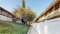 This two-story residential building presents a classic, functional design characterized by a tan stucco exterior with dark brown trim accents. Each level features a covered walkway lined with iron railings, providing balcony-style access to individual units. Exterior staircases connect the ground and upper floors, offering convenient access throughout the building. White vinyl privacy fencing runs along the perimeter, clearly defining the property boundary and providing residents with a sense of seclusion. A paved walkway runs alongside the building, framed by the white fencing on one side and a gently sloping, landscaped hillside on the other. The grounds are generously adorned with mature trees — including tall cypress and broad deciduous varieties — that provide ample shade and greenery throughout the property. Low-growing ground cover blankets the sloped areas, contributing to a well-maintained and verdant outdoor environment. Vintage-style lamp posts are positioned along the walkway, adding a charming detail to the exterior setting. The overall ambiance of the property is one of established, comfortable residential living, with a balance of practical architecture and natural landscaping that creates an inviting and cohesive outdoor environment.
