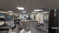 This fitness center is a comprehensively equipped exercise facility designed to support a full range of workout routines. The space is spread across a single floor and organized into distinct activity zones, allowing users to move efficiently between cardio and strength training areas.

The cardio section features a row of Life Fitness treadmills, elliptical trainers, and stationary bikes, each equipped with individual monitors. Large mirrored walls line multiple sections of the facility, visually expanding the space and allowing users to monitor their form. Recessed fluorescent panel lighting set within a drop-tile ceiling provides consistent, even illumination throughout.

The strength training area is well-stocked with cable machines, multi-function weight stations, and a dumbbell rack. Benches and resistance equipment are positioned throughout, offering a variety of targeted workout options. A designated open area near the exit accommodates balance and functional training equipment, including a stability ball machine.

Dark rubber flooring covers the entire facility, providing durability and traction underfoot. Windows with decorative frosted glazing allow natural light to filter in without compromising privacy. The overall layout is practical and efficient, with clear sightlines across the floor supported by strategically placed mirrors.