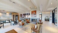 Located in Scottsdale, Arizona, this thoughtfully designed community lounge offers a sophisticated and welcoming environment spread across a generously sized single floor. The open-plan layout is anchored by a dramatic floor-to-ceiling fluted fireplace surround, which serves as the visual centerpiece of the space and is adorned with a bold, warm-toned artwork above. Soaring ceilings with exposed beams amplify the sense of volume throughout.

The lounge is organized into distinct yet seamlessly connected zones. A comfortable seating area features a curated mix of leather and upholstered armchairs arranged on a patterned area rug, ideal for relaxed conversation. Adjacent to this, a dining area is furnished with sleek marble-top round tables and cane-back chairs, bathed in natural light from expansive floor-to-ceiling windows that frame views of the surrounding Scottsdale landscape.

A bar and café area is equipped with counter seating, beverage refrigerators, and warm wood cabinetry, lending a hospitality-forward feel. Large-format light tile flooring unifies the entire space with a clean, contemporary aesthetic.

A sculptural staircase with a curved black metal railing is flanked by a built-in cushioned bench and a lush indoor garden of tropical plants, creating a vibrant and inviting transition point within the space.