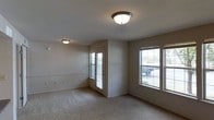 This single-floor space presents a straightforward and functional layout centered around a generously sized living area. The room is finished with wall-to-wall carpet in a neutral tone, contributing to a warm and understated aesthetic that pairs well with the light beige walls throughout. Two flush-mount ceiling light fixtures provide consistent overhead illumination, ensuring the space remains well-lit regardless of the time of day.

Natural light is a notable feature of this living area, with multiple windows positioned along the walls, including a larger double window that offers views of the surrounding outdoor environment. A dedicated exterior door provides direct outdoor access, adding convenience and enhancing the flow of natural light into the space. The windows are fitted with horizontal blinds, allowing for flexible light and privacy control.

The textured ceiling and clean wall lines give the space a simple, no-frills character. The neutral color palette of the carpet, walls, and trim creates a cohesive and versatile backdrop, making the space adaptable to a variety of interior styles and furnishing choices. Overall, the space offers a clean, move-in-ready environment with practical features throughout.