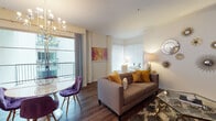 This well-appointed single-floor residence at 11049 Magnolia Blvd in North Hollywood offers a thoughtfully designed layout with distinct living, dining, sleeping, and kitchen areas. The open-concept living room is a visual highlight, featuring rich hardwood-style flooring, a corner bay window with white blinds, and large sliding glass doors that lead to a private balcony, filling the space with natural light. A bold purple accent wall anchors one end of the living area, adorned with an eclectic sunburst mirror gallery and a wall-mounted television, while warm gold and taupe furnishings add a cohesive, contemporary aesthetic. The dining area flows seamlessly from the living space, enhanced by a modern chandelier. The kitchen is equipped with dark espresso cabinetry, stainless steel appliances including a gas range and refrigerator, and granite-style countertops. Two bedrooms offer distinct personalities — one features a cozy, bohemian-inspired design with warm pendant lighting, while the other presents a refined, serene atmosphere. The bathroom includes a combined tub and shower with sliding glass doors and wood-look tile flooring throughout.