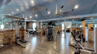This single-floor fitness space combines functional design with distinctive aesthetic character. The most striking architectural feature is the ceiling, which is painted with a detailed sky-and-clouds mural in soft blues and whites, creating an airy, open atmosphere. A large, industrial-style ceiling fan with wide extended blades anchors the center of the room, ensuring effective air circulation throughout.

The walls are accented with rustic stone pillar and archway detailing, adding warmth and texture that contrasts pleasantly with the otherwise modern equipment. Full-length mirrors line multiple walls, visually expanding the space and providing users with proper form-checking capability during workouts.

The flooring consists of durable wood-style planking, offering both aesthetic appeal and practical resilience underfoot. Recessed lighting supplements natural illumination, keeping the environment bright and well-lit.

The facility is equipped with a comprehensive range of exercise machines, including treadmills, cable and resistance machines, and free-weight benches, all arranged to allow comfortable circulation. Motivational signage is incorporated into the mirrored walls, reinforcing the space's energetic and purposeful atmosphere.