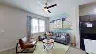 This single-floor residence showcases a cohesive contemporary design with a vibrant, curated aesthetic throughout. The living room serves as a focal point, featuring a ceiling fan with light fixture, light wood-style flooring, and a striking accent wall adorned with horizontal wooden slats in shades of teal, navy, and yellow-green. Natural light filters in through windows dressed with patterned curtains, illuminating the tufted gray sectional sofa, leather accent chair, and geometric round coffee table set atop a soft area rug.

The kitchen is well-appointed with dark espresso cabinetry, a white-topped island with seating for two highlighted by bold yellow stools, pendant lighting overhead, and black appliances including a refrigerator, range, and microwave. Light wood-style flooring extends seamlessly throughout.

A cozy dining nook accommodates a round pedestal table flanked by navy velvet chairs, with colorful abstract artwork adorning the walls above.

The bedroom features a tall channeled blue velvet headboard, ceiling fan, wood-style flooring, and windows with patterned curtains providing natural light. A tufted bench and warm wood nightstands complete the space.

The bathroom includes a full soaking tub with shower, wood-look flooring, and dark cabinetry for storage.
