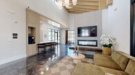 This contemporary single-floor space makes a striking architectural statement with its soaring vaulted ceilings accented by exposed natural wood beam detailing, creating a dramatic sense of openness and volume. Throughout the main area, richly polished concrete floors with a deep, marbled finish reflect light beautifully, anchoring the space in an industrial-modern aesthetic.

The open-concept living and lounge area centers around a sleek, full-width linear fireplace set into a textured stone-clad accent wall, topped by a wall-mounted television. A cozy seating arrangement with a sectional sofa rests atop a geometric patterned area rug, while a secondary seating nook features a classic tufted leather sofa beneath a large-scale abstract artwork, framed by warm wood ceiling elements.

The kitchen is thoughtfully designed with flat-panel light wood cabinetry extending floor to ceiling, white quartz countertops, stainless steel appliances including a built-in oven and French door refrigerator, and a central island with bar seating. Pendant lighting adds warmth over both the island and kitchen counter areas.

Large steel-framed windows and glass entry doors draw in abundant natural light, while a fully glazed partition wall encloses an adjacent conference room, maintaining visual connectivity throughout. The space also includes restroom facilities accessible from the main area.