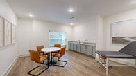 This single-room clinical or wellness space is thoughtfully designed to accommodate both patient consultations and hands-on care. The room is finished with warm-toned luxury vinyl plank flooring throughout, creating a clean and welcoming environment that balances professionalism with comfort.

A key feature of the space is its built-in cabinetry finished in a soft sage green, paired with a light countertop and an integrated sink — ideal for clinical use. Task lighting above the counter ensures a well-lit workspace. Adjacent to the cabinetry, a padded exam table with a mobile stool provides a dedicated area for patient assessment or treatment.

The consultation zone includes a round white table surrounded by tan leather chairs with sleek metal frames, offering a comfortable setting for patient discussions. A large window floods this area with natural light and provides a view of the surrounding greenery, contributing to a calming atmosphere.

The walls are painted in a crisp, neutral white, keeping the space bright and open. Framed botanical and landscape artwork is displayed throughout, softening the clinical aesthetic and adding a refined, welcoming touch. Recessed lighting supplements the natural light, ensuring consistent illumination across the entire room. A dark-framed door provides access to the broader facility.