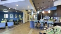 This single-floor space showcases an open-concept design that seamlessly connects a fully equipped kitchen with a social lounge area. The kitchen is outfitted with dark cabinetry, stainless steel appliances, a mosaic tile backsplash, and a large island topped with granite-style countertops, surrounded by cushioned bar stools — ideal for casual dining or socializing. Pendant lighting hangs above the island, adding a warm, modern ambiance complemented by recessed ceiling lights throughout.

The lounge area features bold navy blue accent walls adorned with framed artwork, creating a vibrant yet inviting atmosphere. Seating includes armchairs and bar-height tables, providing flexible arrangements for relaxation or group gatherings. A foosball table visible in the foreground hints at the recreational character of the space.

Columns punctuate the open floor plan, adding architectural interest while maintaining the sense of openness. Large-format tile flooring runs throughout, offering a clean, cohesive look. The combination of contemporary finishes, thoughtful lighting, and multi-functional zones makes this a well-appointed communal space designed for both comfort and engagement.
