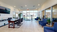 Primrose Villa's single-story clubhouse is designed to serve residents with a range of functional, well-appointed amenities. The main entrance opens into a bright, spacious lobby finished with large-format light tile flooring and floor-to-ceiling glass walls and doors that flood the interior with natural light and offer clear views of the surrounding grounds. A branded welcome mat greets visitors at the entry, while the open layout accommodates a leasing desk with guest seating and a comfortable lounge area furnished with blue accent chairs — ideal for casual meetings or relaxed waiting. A glass-enclosed business center sits adjacent to the lobby, housing multiple computer workstations along a granite-topped counter. The high ceilings throughout the main area create an airy, open atmosphere, accented by a wall-mounted flat-screen display. Moving deeper into the facility, warm hardwood flooring transitions the space into a kitchenette equipped with dark cabinetry, granite countertops, a sink, microwave, and refrigerator. Dual ADA-accessible drinking fountains are conveniently located near the restrooms. The fitness room features rubber flooring and is outfitted with cardio equipment including a treadmill and elliptical, as well as a multi-function cable machine and stationary bike, with natural light provided by horizontal windows.