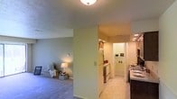 This single-floor residence offers a practical and straightforward layout that integrates living and functional spaces seamlessly. The living area is finished with wall-to-wall carpeting and painted in a soft, neutral green-gray tone, creating a calm and understated ambiance. A flush-mount ceiling light provides overhead illumination, while a table lamp in the corner adds warm accent lighting. The space accommodates comfortable furnishings and offers a relaxed, livable atmosphere.

A central dividing wall separates the living area from the kitchen, which features tile flooring, wood-toned upper cabinetry, a white refrigerator, and a white range. Recessed lighting in the kitchen provides bright, even illumination suited for meal preparation. The kitchen connects directly to an adjoining utility or laundry area, which shares the same tile flooring and benefits from additional recessed lighting, making it a well-lit and functional support space.

A countertop or bar surface is partially visible along the right side, suggesting a transitional space between the kitchen and living areas. The textured ceilings are consistent throughout, tying the spaces together with a cohesive finish. Overall, the residence is designed for efficient, comfortable single-floor living.