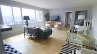 This single-floor space showcases a thoughtfully designed interior with a welcoming and functional layout. The living area serves as the heart of the space, featuring warm light-toned tile flooring that extends throughout, creating a sense of continuity and openness. Large windows flood the room with natural light and frame expansive outdoor views, adding an airy, elevated feel to the interior.

The living area is styled with mid-century modern furnishings, including a teal upholstered sofa, paired accent chairs in soft white with contrasting orange cushions, and a natural wood media console supporting a flat-screen television. A glass-top console table along the window wall, adorned with a statement table lamp and decorative accessories, adds a refined touch. A bold black-and-white striped area rug anchors the dining area, where a glass dining table is partially visible, complementing the overall contemporary aesthetic.

A tall potted plant adds a natural, organic element to the corner of the room, while neutral grey walls provide a clean, versatile backdrop throughout. A glimpse through an open doorway hints at additional rooms beyond, suggesting a well-connected and practical floor plan. Overall, the space exudes a bright, modern ambiance with a balanced blend of comfort and style.