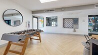 Located at 4722 E Bell Rd in Phoenix, Arizona, this single-floor community lounge is a well-appointed social space designed for both relaxation and recreation. The room features striking herringbone-patterned light hardwood flooring that runs throughout, lending a clean, contemporary aesthetic to the entire space.

The lounge is divided into distinct functional zones. A comfortable seating area anchors one side of the room, furnished with a tan leather sofa, a sculptural upholstered armchair, and a round wooden coffee table set atop a teal patterned area rug. Bold teal accent walls provide a vibrant contrast to the bright white walls and ceiling, while colorful modern artwork adds personality throughout. A wall-mounted flat-screen television and a sleek electric fireplace serve as a focal point in the lounge area.

The recreational zone features a full-size foosball table alongside a multi-seat dining or game table, making it ideal for group gatherings. A large round mirror and an oversized wall-mounted board game add playful, decorative touches. Recessed and flush-mount ceiling lighting, along with multiple windows and glass double doors, ensures the space is bright and well-lit. Views into an adjacent fitness area are visible through interior windows, highlighting the broader amenity offerings of this community.