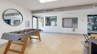 Located at 4722 E Bell Rd in Phoenix, Arizona, this single-floor community lounge is a well-appointed social space designed for both relaxation and recreation. The room features striking herringbone-patterned light hardwood flooring that runs throughout, lending a clean, contemporary aesthetic to the entire space.

The lounge is divided into distinct functional zones. A comfortable seating area anchors one side of the room, furnished with a tan leather sofa, a sculptural upholstered armchair, and a round wooden coffee table set atop a teal patterned area rug. Bold teal accent walls provide a vibrant contrast to the bright white walls and ceiling, while colorful modern artwork adds personality throughout. A wall-mounted flat-screen television and a sleek electric fireplace serve as a focal point in the lounge area.

The recreational zone features a full-size foosball table alongside a multi-seat dining or game table, making it ideal for group gatherings. A large round mirror and an oversized wall-mounted board game add playful, decorative touches. Recessed and flush-mount ceiling lighting, along with multiple windows and glass double doors, ensures the space is bright and well-lit. Views into an adjacent fitness area are visible through interior windows, highlighting the broader amenity offerings of this community.