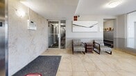 Located at 221 Glenridge Ave in St. Catharines, Ontario, this building lobby serves as the welcoming entrance to a Homestead residential property. The ground-floor space is finished with light beige ceramic tile flooring accented by grey carpet runners, creating a clean and functional aesthetic throughout.

Upon entry, floor-to-ceiling glass panels and automatic doors flood the lobby with natural light while offering clear sightlines to the exterior parking area. Flush-mounted ceiling light fixtures provide consistent ambient illumination across the space.

Moving inward, a pair of stainless steel elevators set against a stone-textured wall serve as the primary vertical access point for residents. A resident information board is mounted nearby, offering building notices and updates. Adjacent to the elevators, a glass-panel door provides access to the main residential corridor.

A comfortable seating area is positioned toward the interior, furnished with two upholstered armchairs and a small side table, alongside a framed abstract artwork that adds a touch of character to the neutral walls. A wooden bench near the elevator bank offers additional seating. Security is supported by a visible surveillance camera.

The lobby also connects to a secondary corridor, with a mailbox area visible through a side passage. Overall, the space presents a practical, well-maintained, and welcoming environment for residents and visitors alike.