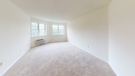 Located at 601 Riverside Ave in Lyndhurst, NJ, Unit 220 is a single-floor residence that makes efficient use of its space with a practical and comfortable layout. The kitchen is well-appointed with warm honey-toned wood cabinetry extending to the ceiling, offering generous storage throughout. Light-colored countertops provide ample workspace, while tile flooring adds durability to the area. White appliances, including a dishwasher, range, and refrigerator, are included. The living area is generously sized, featuring plush neutral-toned carpeting and crisp white walls that lend a clean, bright ambiance. A pair of windows allows natural light to flow in, offering views of surrounding greenery. A wall-mounted HVAC unit provides climate control. The bedroom mirrors this bright, open feel with its own window framing outdoor views and matching carpet flooring, creating a calm and restful environment. The bathroom is finished with tile surrounding a full soaking tub and shower combination, complemented by a single-sink vanity with wood cabinetry and a lighted medicine cabinet mirror above. Clean white walls and neutral finishes throughout the unit create a cohesive, move-in ready aesthetic.