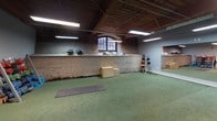 Located at 300 S Firestone St in Gastonia, North Carolina, this fitness facility occupies a single-floor space rich in industrial character and thoughtfully outfitted for a comprehensive training experience. The building's heritage is evident throughout, with exposed brick walls, warm wood-paneled ceilings, visible ductwork, and sturdy structural columns that lend the space a distinctive, converted-warehouse aesthetic.

The facility is divided into clearly defined training zones. A functional training area features green turf flooring and is stocked with kettlebells, dumbbells on a multi-tier rack, balance domes, medicine balls, plyo boxes, resistance bands, and yoga mats — accommodating a wide range of movement-based and accessory workouts. A large wall-mounted mirror enhances spatial awareness and form-checking in this zone.

The strength training area transitions to dark rubber flooring and houses power racks, barbells, weight plates, a sled machine, and a treadmill, providing a robust setup for heavy lifting and conditioning. Full-length mirrors line portions of this section as well, maximizing functionality.

Arched industrial windows allow natural light to filter into the turf zone, complementing the overhead fluorescent lighting that illuminates the entire floor. Storage cubbies and organized equipment racks keep the space tidy and accessible. The layout flows openly across zones, making navigation intuitive for users of all training styles.