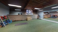 Located at 300 S Firestone St in Gastonia, North Carolina, this fitness facility occupies a single-floor space rich in industrial character and thoughtfully outfitted for a comprehensive training experience. The building's heritage is evident throughout, with exposed brick walls, warm wood-paneled ceilings, visible ductwork, and sturdy structural columns that lend the space a distinctive, converted-warehouse aesthetic.

The facility is divided into clearly defined training zones. A functional training area features green turf flooring and is stocked with kettlebells, dumbbells on a multi-tier rack, balance domes, medicine balls, plyo boxes, resistance bands, and yoga mats — accommodating a wide range of movement-based and accessory workouts. A large wall-mounted mirror enhances spatial awareness and form-checking in this zone.

The strength training area transitions to dark rubber flooring and houses power racks, barbells, weight plates, a sled machine, and a treadmill, providing a robust setup for heavy lifting and conditioning. Full-length mirrors line portions of this section as well, maximizing functionality.

Arched industrial windows allow natural light to filter into the turf zone, complementing the overhead fluorescent lighting that illuminates the entire floor. Storage cubbies and organized equipment racks keep the space tidy and accessible. The layout flows openly across zones, making navigation intuitive for users of all training styles.