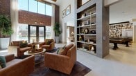 This single-floor communal amenity space is thoughtfully designed to accommodate a variety of social, recreational, and professional needs. The main lounge area serves as the centerpiece, featuring exposed brick accent walls, large windows that invite natural light, and wide-format tile flooring. Clusters of plush seating—including green velvet ottomans, neutral sectional sofas, and cognac leather armchairs—are arranged around circular coffee tables beneath a striking cluster of globe pendant lights in warm gold tones.

A built-in shelving unit with wood-paneled backing provides decorative display space, while a tile-surround linear fireplace with a mounted television above anchors a key focal point in the room. Structural columns clad in natural wood add warmth throughout the corridor leading deeper into the space.

The game area includes a billiards table alongside a casual dining setup adjacent to exposed brick and exterior-facing windows. A fully equipped kitchen bar features a dark stone countertop island with seating, stainless steel appliances, pendant lighting, and a bold geometric tile backsplash beneath a textured ceiling detail.

A glass-walled conference room enclosed by black steel-framed sliding doors provides a semi-private professional workspace with a conference table, ergonomic seating, and a wall-mounted display screen.