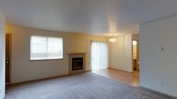 This two-floor residence offers a thoughtfully designed layout across its levels, providing ample room for everyday living. The main living area serves as the heart of the home, featuring a built-in fireplace that anchors the space and adds warmth and character. Large windows fitted with blinds allow natural light to filter in, while a sliding glass door with vertical blinds provides access to the outdoors and enhances the sense of openness. An elegant chandelier adds a refined touch to the living space, complementing the neutral tones of the walls and light hardwood-style flooring. Plush carpeting in the main living area offers comfort underfoot, creating a cozy atmosphere. The smooth, high ceiling contributes to the airy feel of the interior. Transitioning through the space, an adjacent area hints at additional rooms beyond, suggesting a well-connected floor plan. The overall aesthetic is clean and neutral, offering a versatile canvas that suits a variety of interior styles. With its combination of natural light, classic fixtures, and practical features spread across two floors, this residence provides a balanced and livable environment.