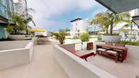 Located at Harbour Island in Oxnard, CA, this single-level outdoor space embodies the relaxed coastal lifestyle of Southern California. The generously sized private patio is finished with large-format neutral stone tiles that lend a clean, contemporary aesthetic. Comfortable teak wood outdoor furniture, including a dining table, benches, and lounge chairs with red cushions, provide inviting areas for both dining and relaxation beneath a covered overhang.

The patio is framed by crisp white stucco walls and raised planters filled with lush tropical greenery, including bamboo and ornamental grasses, which add privacy and a resort-like ambiance. Sliding glass doors connect the outdoor space directly to the interior, allowing for a seamless indoor-outdoor flow bathed in natural California sunlight.

The surrounding Harbour Island community enhances the setting with a beautifully landscaped shared courtyard featuring additional seating areas shaded by bold yellow umbrellas, tall palms, and well-maintained garden beds. Contemporary multi-story architecture with clean lines and large windows characterizes the neighboring buildings, contributing to a cohesive, upscale coastal community aesthetic. The open sky and proximity to the Pacific coast make this an exceptional outdoor retreat.