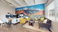 This single-story clubhouse offers a well-designed, open-concept layout that seamlessly blends functionality with contemporary style. The space is defined by soaring vaulted ceilings with exposed white-painted beams and track lighting, creating a bright and airy atmosphere throughout.

Large transom windows line the upper walls, flooding the interior with abundant natural light and enhancing the sense of openness. Warm hardwood flooring runs continuously across the entire space, adding a cohesive and inviting feel underfoot.

The kitchen area features a substantial granite-topped peninsula with modern wire bar stools, dark wood cabinetry, and ample counter space — ideal for community gatherings and events. Adjacent to the kitchen, a spacious open lounge area serves as the social heart of the clubhouse, anchored by a dramatic floor-to-ceiling landscape mural depicting a vibrant mountain scene, adding a bold visual focal point to the room. Comfortable seating, including accent chairs and a sofa, creates a relaxed and welcoming environment.

A dedicated workspace with a computer station provides residents with a practical area for everyday tasks. The clubhouse also includes a coat area equipped with a sliding barn door and a coat rack, adding a practical yet stylish entryway detail. The overall ambiance is modern, bright, and community-oriented.