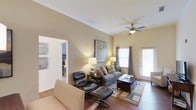 Located at 115 Amberleigh Dr in Wilmington, North Carolina, this single-floor residence offers a comfortable and thoughtfully designed living space. The living room serves as the heart of the home, featuring warm neutral-toned walls that create a welcoming ambiance throughout. Hardwood flooring adds a classic touch underfoot, complemented by a patterned area rug that anchors the seating arrangement. A ceiling fan with an integrated light fixture provides both comfort and illumination, while natural light streams in through windows dressed with elegant curtain panels. The living area is well-proportioned, accommodating a full sofa, a classic lounge chair with matching ottoman, and a wooden coffee table — offering both style and functionality. A decorative art piece on the wall adds a pop of color and personality to the space. An open doorway connects the living room to an adjacent bedroom, enhancing the sense of flow and accessibility throughout the home. The layout is practical and open, making it easy to move between spaces. Overall, this residence combines a relaxed, residential charm with tasteful design elements that make it a comfortable and livable home in the Wilmington area.