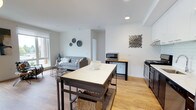 This single-floor space showcases an open-concept layout that connects the kitchen, dining, and living areas into one cohesive, functional environment. The interior is finished with warm hardwood-style flooring that runs throughout, complementing the clean white walls and creating a bright, welcoming atmosphere.

The kitchen is outfitted with stainless steel appliances, including a gas range, and features ample cabinetry in white tones with light wood countertops. A central island with a white countertop and wire basket storage beneath serves as both a workspace and an informal dining area, adding versatility to the space.

Adjacent to the kitchen, a dedicated dining or utility counter hosts a stainless steel microwave and additional storage, bridging the transition to the living area. The living space is defined by a contemporary gray sofa, decorative wall accents, and a window that draws in natural light.

Recessed lighting in the ceiling provides consistent illumination throughout. The overall design aesthetic is modern and understated, with industrial-inspired touches balanced by soft, neutral tones that give the space a clean and comfortable feel.