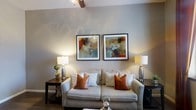 This single-floor space offers a comfortable and thoughtfully curated living environment. The living area serves as the heart of the home, centered around a plush, light-toned sofa accented with warm terracotta and soft striped throw pillows, creating an inviting and relaxed atmosphere. A pair of framed abstract artworks with rich earthy tones — deep reds, burnt oranges, and muted teals — are displayed symmetrically above the sofa, adding a sophisticated artistic focal point to the room. Flanking the seating area are matching decorative table lamps with textured silver bases, casting a warm and ambient glow that enhances the overall coziness of the space. The walls are finished in a soft, neutral greige tone that complements the furnishings and contributes to a calm, harmonious aesthetic. A ceiling fan is visible above, offering both functional air circulation and a casual design touch. The overall ambiance of the space is warm, polished, and livable — balancing decorative detail with everyday comfort. The neutral foundation throughout allows for versatile styling, making the space feel both timeless and welcoming to all who enter.