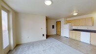 This single-floor unit offers a straightforward and functional layout designed to maximize everyday comfort. The living area is finished with soft wall-to-wall carpeting, creating a warm and inviting atmosphere, while neutral white walls provide a clean, versatile backdrop throughout the space. Flush-mount ceiling lighting illuminates the room evenly, contributing to a bright and practical interior.

The living area flows naturally toward the entryway, which features a solid wood front door that adds a touch of warmth to the otherwise neutral palette. A wall-mounted heating unit is conveniently positioned in the living space, ensuring efficient climate control.

Transitioning into the kitchen area, the flooring shifts to a tile surface, clearly defining the cooking and dining zone. Light wood cabinetry lines the upper walls, offering ample storage, while dark countertops provide a subtle contrast. A full-size white refrigerator anchors the kitchen, and the overall layout is compact yet well-organized for everyday meal preparation.

A hallway visible from the main living area leads to additional rooms, suggesting a practical division between living and private spaces. The textured ceiling throughout adds a subtle architectural detail, and the overall design of this unit prioritizes clean lines, functional storage, and a cohesive, understated aesthetic.