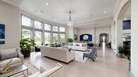 Located at 8335 Narcoossee Rd in Orlando, FL, this single-floor amenity center offers a well-rounded collection of spaces designed for comfort, productivity, and fitness. The expansive living room serves as the heart of the facility, boasting soaring ceilings, a dramatic curved wall of floor-to-ceiling windows, and an abundance of natural light. Elegant furnishings in neutral tones, a statement chandelier, crown molding, and lush indoor plants create a sophisticated yet welcoming atmosphere with views of the surrounding Florida landscape. An adjacent dining area features a live-edge wood table with navy blue chairs, coastal-inspired artwork, and a built-in beverage station framed by a decorative arch. The connected lounge area offers additional seating, a wall-mounted TV, and arched architectural details throughout. The business center provides multiple workstations with granite countertops, a built-in bench seating area, and marble-top café tables. Two dedicated exercise rooms cater to diverse fitness needs — one equipped with treadmills, cable machines, and free weights beneath soaring ceilings with large windows, and the other featuring spin bikes, rowing machines, floor-to-ceiling mirrors, and acoustic panels. A covered outdoor lounge with wicker seating and tropical landscaping completes this comprehensive amenity offering.
