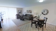 This single-floor space features a seamlessly integrated open-concept layout that combines a living area and dining space into one cohesive environment. The living area is anchored by a neutral-toned sofa and matching armchair arranged around a round coffee table atop a patterned area rug, creating a comfortable and inviting seating arrangement. Decorative wall art adds visual interest to the soft, light-coloured walls, while a pair of table lamps provide warm ambient lighting to complement the flush ceiling light fixture overhead.

The dining area flows naturally from the living space and is furnished with a dark-toned round table surrounded by matching chairs, making it well-suited for everyday meals or entertaining. A decorative oversized clock on the adjacent wall serves as a bold focal point in this zone.

Large windows along the perimeter of the living area allow generous amounts of natural light to filter in, contributing to the bright and airy feel of the space. The flooring transitions between a classic parquet hardwood and the area rug, adding texture and warmth underfoot. Overall, the space maintains a clean, neutral aesthetic that feels both functional and welcoming.