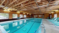 This single-story recreational facility is defined by two primary spaces: a generously sized indoor lap pool area and an adjoining fitness room. The pool hall is the architectural centerpiece of the facility, showcasing warm wood-paneled walls and soaring exposed timber roof trusses that lend the space a rustic, lodge-like character. The pool itself features multiple lap lanes and is bathed in natural light from a row of large windows lining one wall, offering views of the surrounding greenery. Poolside seating with teal-colored chairs provides a comfortable area for relaxation. A glass partition separates the pool area from the fitness room, maintaining visual connectivity between the two spaces. The fitness room offers a well-equipped workout environment, featuring treadmills, a Body-Solid multi-station strength training machine, and additional cardio equipment. Floor-to-ceiling mirrors amplify the sense of space and assist with exercise form. The fitness room is finished with a drop tile ceiling, carpet flooring, and large windows that bring in natural light and views of the outdoor landscape. Together, the two spaces create a comprehensive, self-contained recreational amenity suited for both aquatic exercise and cardiovascular and strength training.