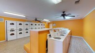 The Clothes Care Center is a well-equipped, single-floor laundromat designed to accommodate a high volume of laundry needs efficiently. The space is immediately recognizable by its bold, vibrant orange walls and clean white trim, creating a bright and energetic atmosphere throughout. Light wood-look flooring runs across the entire facility, contributing to a tidy and modern feel.

The main area is lined with multiple top-loading washing machines arranged along the perimeter and around a central island structure, providing ample washing capacity. A substantial bank of stacked dryer units occupies one wall, offering equally generous drying capacity. Each machine is clearly branded and equipped with operating instructions posted visibly for customer convenience.

Overhead, multiple ceiling fans are evenly distributed across the space, ensuring consistent air circulation and comfort for patrons. Fluorescent lighting fixtures provide bright, even illumination throughout. A folding table positioned near a window offers a practical surface for sorting and folding laundry, with natural light streaming in through the windows and views of outdoor greenery.

Additional amenities include a bulletin board for notices, a fire extinguisher for safety compliance, a waste bin, and clearly posted security and operating notices. The layout is open and spacious, allowing for easy movement between machines. The Clothes Care Center is a clean, functional, and well-maintained facility designed to serve its customers effectively.