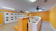 The Clothes Care Center is a well-equipped, single-floor laundromat designed to accommodate a high volume of laundry needs efficiently. The space is immediately recognizable by its bold, vibrant orange walls and clean white trim, creating a bright and energetic atmosphere throughout. Light wood-look flooring runs across the entire facility, contributing to a tidy and modern feel.

The main area is lined with multiple top-loading washing machines arranged along the perimeter and around a central island structure, providing ample washing capacity. A substantial bank of stacked dryer units occupies one wall, offering equally generous drying capacity. Each machine is clearly branded and equipped with operating instructions posted visibly for customer convenience.

Overhead, multiple ceiling fans are evenly distributed across the space, ensuring consistent air circulation and comfort for patrons. Fluorescent lighting fixtures provide bright, even illumination throughout. A folding table positioned near a window offers a practical surface for sorting and folding laundry, with natural light streaming in through the windows and views of outdoor greenery.

Additional amenities include a bulletin board for notices, a fire extinguisher for safety compliance, a waste bin, and clearly posted security and operating notices. The layout is open and spacious, allowing for easy movement between machines. The Clothes Care Center is a clean, functional, and well-maintained facility designed to serve its customers effectively.