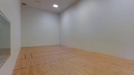 This indoor racquetball court is a dedicated, single-floor athletic facility designed for competitive and recreational play. The court features a classic light-toned hardwood floor, finished to a smooth surface and marked with the standard red service and receiving lines that define regulation play. The spacious playing area offers ample room for full court movement, giving players the freedom to engage in dynamic, fast-paced rallies.

All four walls are finished in a clean, bright white, a standard characteristic of racquetball courts that maximizes visibility of the ball during play. The smooth, uninterrupted wall surfaces extend from floor to ceiling without obstructions, ensuring consistent ball rebound across every angle. The high ceiling contributes to the open, airy feel of the space while accommodating the full vertical range of play.

Overhead flush-mounted lighting fixtures provide even, well-distributed illumination throughout the court, minimizing shadows and ensuring clear sightlines for players. The overall environment is clean, minimalist, and purpose-built — every element of the space is oriented toward functionality and an optimal playing experience. This court is well-suited for individual practice, one-on-one matches, or doubles play.