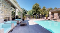 This single-story property showcases a well-appointed outdoor pool area designed for relaxation and leisure. The building exterior features classic shingle siding in a soft teal-green hue, accented by crisp white trim and large floor-to-ceiling glass doors and windows that allow natural light to flow freely between the interior and the pool deck. A stone accent wall adds a touch of textural contrast to the façade, lending the space a refined, welcoming character.

The expansive concrete pool deck provides generous space for lounging and entertaining, centered around a clear, light-blue swimming pool enclosed by a wrought iron security fence with a gated entry. A comfortable seating arrangement consisting of wicker chairs and a low coffee table with bright aqua cushions is positioned along the building's covered edge, offering a shaded retreat that overlooks the pool.

Lush mature trees surround the perimeter, providing natural privacy and a verdant backdrop under open skies. Potted flowering plants, including vibrant petunias and ornamental tropicals, are thoughtfully placed near the building's entrance, adding splashes of color. A decorative gazebo is visible in the background, further enhancing the outdoor amenity offerings of this inviting space.