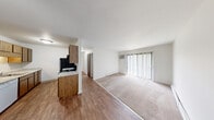 This single-floor, 3-bedroom, 1-bathroom unit offers a practical and efficient layout designed to maximize functionality. The open-concept living and kitchen area forms the heart of the home, with wood-look flooring in the kitchen transitioning to carpet in the living space. The kitchen is equipped with wood-toned cabinetry, granite-look countertops, black appliances including a range, microwave, and refrigerator, along with a ceiling fan and convenient built-in open shelving for additional storage. A large sliding glass door in the living area fills the space with natural light and is complemented by vertical blinds for privacy. A wall-mounted air conditioning unit and baseboard heating ensure year-round comfort throughout. The three bedrooms are carpeted and offer a quiet, neutral palette, with the primary bedroom featuring its own wall A/C unit. Each bedroom includes closet space, with one closet offering a full hanging rod and shelf. The single bathroom features a wood-toned vanity with a white countertop, a large mirror, wood-look flooring, and a baseboard heater. Overall, the unit presents a clean, straightforward living environment with practical finishes throughout.