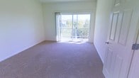 This single-floor space presents a clean, minimalist interior characterized by smooth white walls and wall-to-wall carpeting in a neutral tone. The room is generously proportioned, offering an open feel with unobstructed floor space throughout. A standout feature is the large sliding glass door positioned along the far wall, framed by simple vertical drapes that allow an abundance of natural light to fill the interior. The door provides direct access to a balcony enclosed by a white railing, beyond which a scenic view of trees and a residential landscape is visible, adding a sense of depth and connection to the outdoors. The white baseboards provide a clean visual contrast against the beige carpeting, contributing to the room's bright, airy atmosphere. The overall design is straightforward and functional, with a neutral color palette that creates a versatile canvas. The combination of natural light, outdoor access, and elevated views gives the space a pleasant, open ambiance well-suited to a variety of uses.