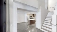 This two-story space showcases a contemporary design defined by clean lines, an open layout, and an impressive sense of vertical space. The main level features polished concrete floors that reflect light throughout, contributing to the modern industrial aesthetic that runs through the entire space.

The kitchen is a standout feature, equipped with stainless steel appliances including a refrigerator, range, and microwave. Warm wood-toned cabinetry lines the walls, providing ample storage, while a central island with a light-colored countertop offers additional workspace and a natural gathering point. Recessed lighting illuminates the kitchen efficiently.

The open living area benefits from soaring double-height ceilings, creating an expansive, airy atmosphere on the main floor. Crisp white walls contrast with the warm tones of the kitchen cabinetry and the cool sheen of the concrete floors. A staircase with clean, open treads leads to the second floor, adding architectural interest while maintaining the space's minimalist character. Overall, the design balances industrial materials with warm finishes for a refined, contemporary feel.