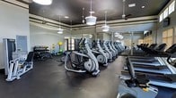 This fitness center offers a comprehensive workout environment designed to accommodate a wide range of exercise routines. The space is anchored by a row of elliptical trainers and cardio machines that stretch across the main floor, complemented by a dedicated section of treadmills along the perimeter. The free weights area is well-stocked with a full rack of dumbbells and a colorful assortment of medicine balls and kettlebells, catering to both strength and functional training needs. Large mirrors line the walls, enhancing the sense of openness and allowing users to monitor their form throughout their workouts. The interior features a dark, contrasting ceiling with decorative molding trim along the upper walls, giving the space a polished, upscale aesthetic. Overhead, a combination of globe pendant lights and ceiling fans provides ample illumination while maintaining comfortable air circulation throughout. The flooring consists of durable rubber matting, ideal for high-impact activities and equipment use. Natural light filters in through windows along one wall, adding brightness to the well-lit interior. The overall ambiance is clean, modern, and purposefully laid out to support an effective and motivating fitness experience.