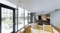 This contemporary single-floor space is defined by its sleek, open-concept design and abundant natural light. The lounge area anchors the space with plush, neutral-toned seating and direct access to an expansive outdoor terrace through full-height glass doors and floor-to-ceiling windows. The glazed facade frames views of a well-maintained outdoor courtyard and the surrounding urban skyline, creating a seamless indoor-outdoor connection.

The interior transitions smoothly into a modern kitchen and dining area, where a large island with bar-height seating serves as both a functional workspace and a social gathering point. Dark cabinetry and a bold range hood provide a striking contrast against the white countertops and bright ceiling, while recessed lighting throughout ensures a well-lit environment at all hours.

Dark hardwood flooring runs consistently across the space, grounding the clean, minimalist aesthetic. The ceilings are smooth and bright white, fitted with recessed lighting and integrated HVAC vents that maintain a tidy, uncluttered appearance. The overall ambiance is modern and sophisticated, balancing openness and warmth in a highly functional layout well-suited for both everyday use and entertaining.