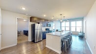 This thoughtfully designed 4-bedroom, 4-bathroom unit offers a functional and modern layout across a single floor. The open-concept kitchen and living area serves as the heart of the space, featuring rich dark wood cabinetry, stainless steel appliances, granite countertops, and a large island with bar seating illuminated by pendant lighting. The adjacent living room benefits from oversized windows with horizontal blinds that frame elevated views of the surrounding neighborhood, while an exposed structural ceiling detail adds an industrial character to the space. Hardwood-style flooring flows throughout the common areas, adding warmth and continuity. Each of the four bedrooms is carpeted and equipped with a bed frame and a dedicated desk and chair workspace, with windows providing natural light and neighborhood views. The four bathrooms are finished consistently, featuring tile flooring, granite-topped vanities, and full tub-shower combinations with chrome fixtures. Two closets provide additional storage throughout the unit. The overall design is clean, neutral, and functional, making it well-suited for shared occupancy. Recessed lighting throughout ensures well-lit common areas, while each private bedroom maintains a quiet, focused atmosphere.