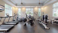 Located at 6450 Mercantile Dr E in Frederick, Maryland, this amenity space at Century Clearbrook offers residents a well-appointed fitness center and a resort-style outdoor pool. The fitness center is housed in a single-story structure with classic architectural detailing, including crown molding, tray ceilings, and large white-shuttered windows that invite generous natural light. The gym is comprehensively equipped with treadmills, elliptical trainers, a cable machine, free weights, a dumbbell rack, and a multi-station strength system, all set on durable rubber flooring. A wall-mounted television and pendant lighting add comfort to the workout environment. Entry to the fitness center is through elegant glass-paneled double French doors beneath a transom window, with a branded Century Clearbrook entry mat welcoming visitors. The outdoor pool area is equally impressive, featuring a generously sized swimming pool with a curved design and clear blue water. A charming stone pavilion with a decorative fireplace serves as a focal point at the far end of the pool deck, which is lined with lounge chairs and accented by potted plants. Mature trees and manicured greenery create a private, park-like setting, with the residential buildings visible in the background.
