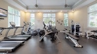 Located at 6450 Mercantile Dr E in Frederick, Maryland, this amenity space at Century Clearbrook offers residents a well-appointed fitness center and a resort-style outdoor pool. The fitness center is housed in a single-story structure with classic architectural detailing, including crown molding, tray ceilings, and large white-shuttered windows that invite generous natural light. The gym is comprehensively equipped with treadmills, elliptical trainers, a cable machine, free weights, a dumbbell rack, and a multi-station strength system, all set on durable rubber flooring. A wall-mounted television and pendant lighting add comfort to the workout environment. Entry to the fitness center is through elegant glass-paneled double French doors beneath a transom window, with a branded Century Clearbrook entry mat welcoming visitors. The outdoor pool area is equally impressive, featuring a generously sized swimming pool with a curved design and clear blue water. A charming stone pavilion with a decorative fireplace serves as a focal point at the far end of the pool deck, which is lined with lounge chairs and accented by potted plants. Mature trees and manicured greenery create a private, park-like setting, with the residential buildings visible in the background.