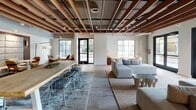 Located at 3200 Paseo Village Way in San Diego, CA, this single-floor amenity space blends contemporary design with warm, inviting finishes throughout. The open layout is anchored by striking exposed wood beam ceilings with integrated track lighting, creating an industrial-modern aesthetic softened by light wood flooring and neutral tones.

The lounge area offers generous seating arrangements, including modular sofas and accent chairs set atop layered area rugs, fostering casual gatherings. A standout nook features a tall upholstered panel wall framing built-in banquette seating alongside woven rattan chairs and bistro-style tables — ideal for intimate conversations or small group meetups.

The bar and kitchen zone is a centerpiece of the space, showcasing marble-topped counters with warm wood cabinetry, a built-in beverage tap, and a mosaic tile backsplash. Smoked glass pendant lights hang above, adding a refined ambiance. Display shelving and neon accent lighting lend a social, lounge-bar feel.

Floor-to-ceiling steel-framed windows flood portions of the space with natural light while offering views of the landscaped outdoor courtyard. Additional seating alcoves near the windows provide a tranquil retreat. Together, these elements create a cohesive, upscale communal environment that balances functionality with sophisticated style.
